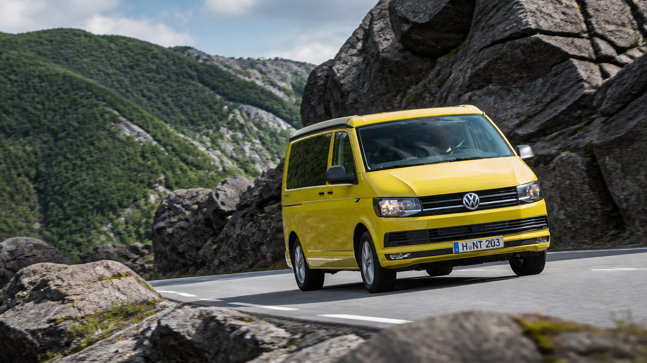 Van Volkswagen amarela dirigindo por estrada sinuosa entre pedras e montanhas verdes.