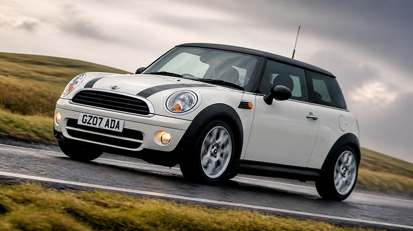 Carro Mini Cooper branco com teto e faixas pretas dirigindo em estrada molhada com céu nublado.